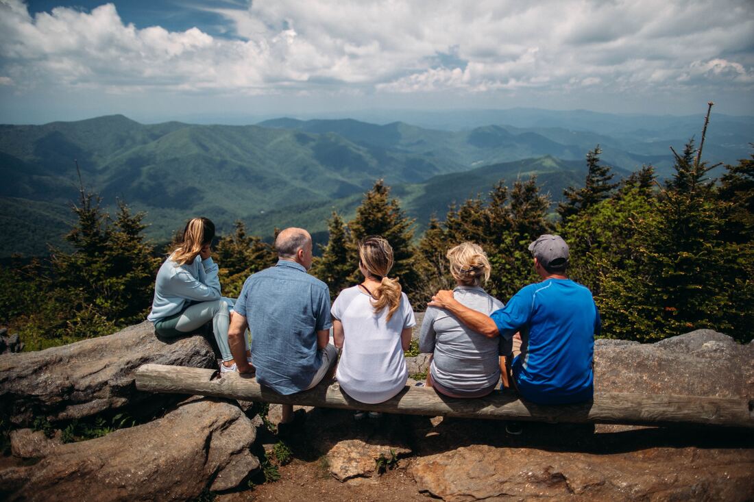 A family of five sits on a hilltop and overlooks views from the mountains while having a conversation.