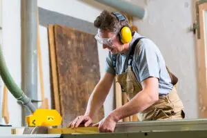 Man wearing earmuffs and safety glasses while operating woodworking equipment – protecting hearing from loud noise exposure to prevent hearing loss.
